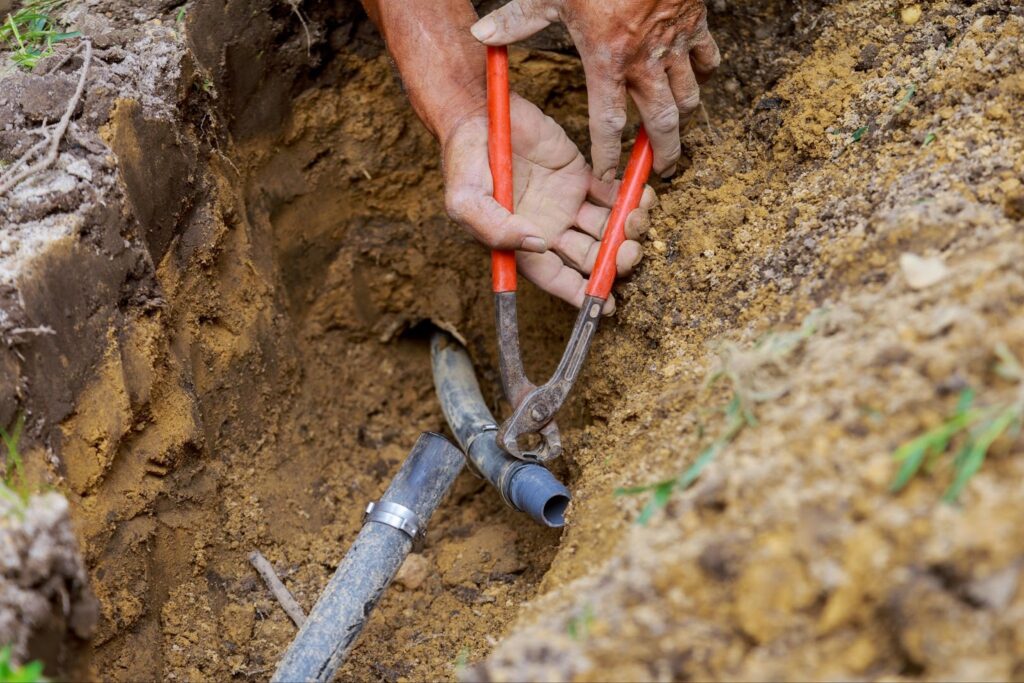A pair of hands using red-handled pliers to repair an underground pipe inside a dirt trench.