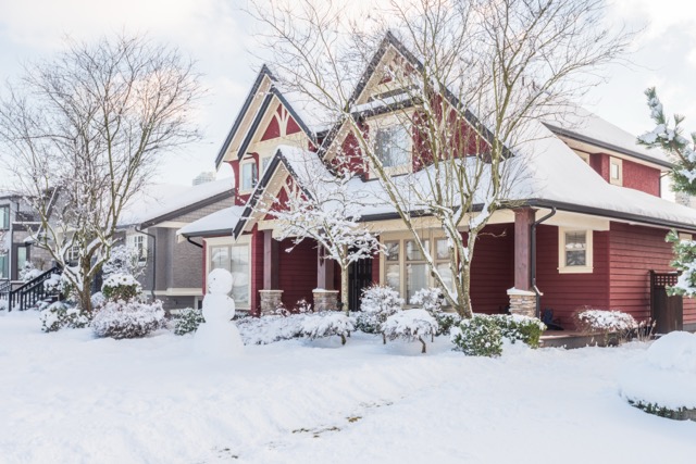 Red house with snow-covered roof, snowy yard, snowman in front, bare trees, winter scene.