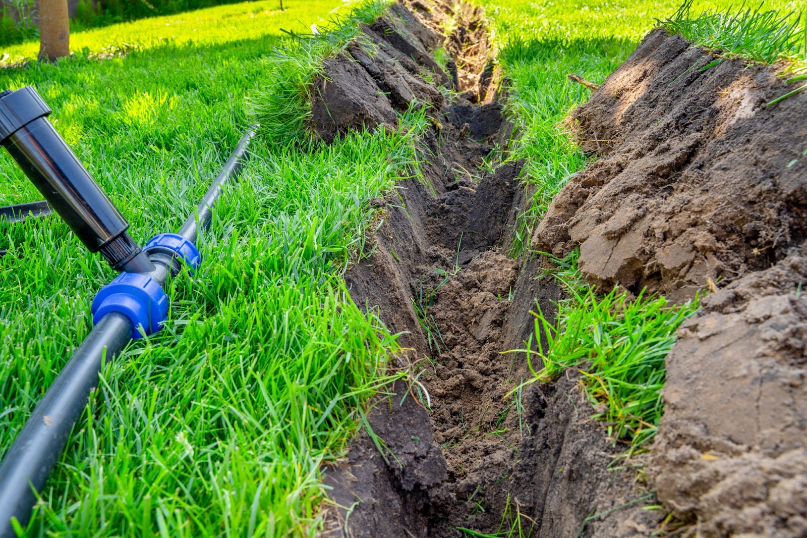 A freshly dug trench in a green lawn for the installation of an underground sprinkler system, with black irrigation pipes and a sprinkler head visible in the foreground.