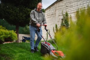 A person mowing a green lawn with a lawnmower near a white brick wall and trees.
