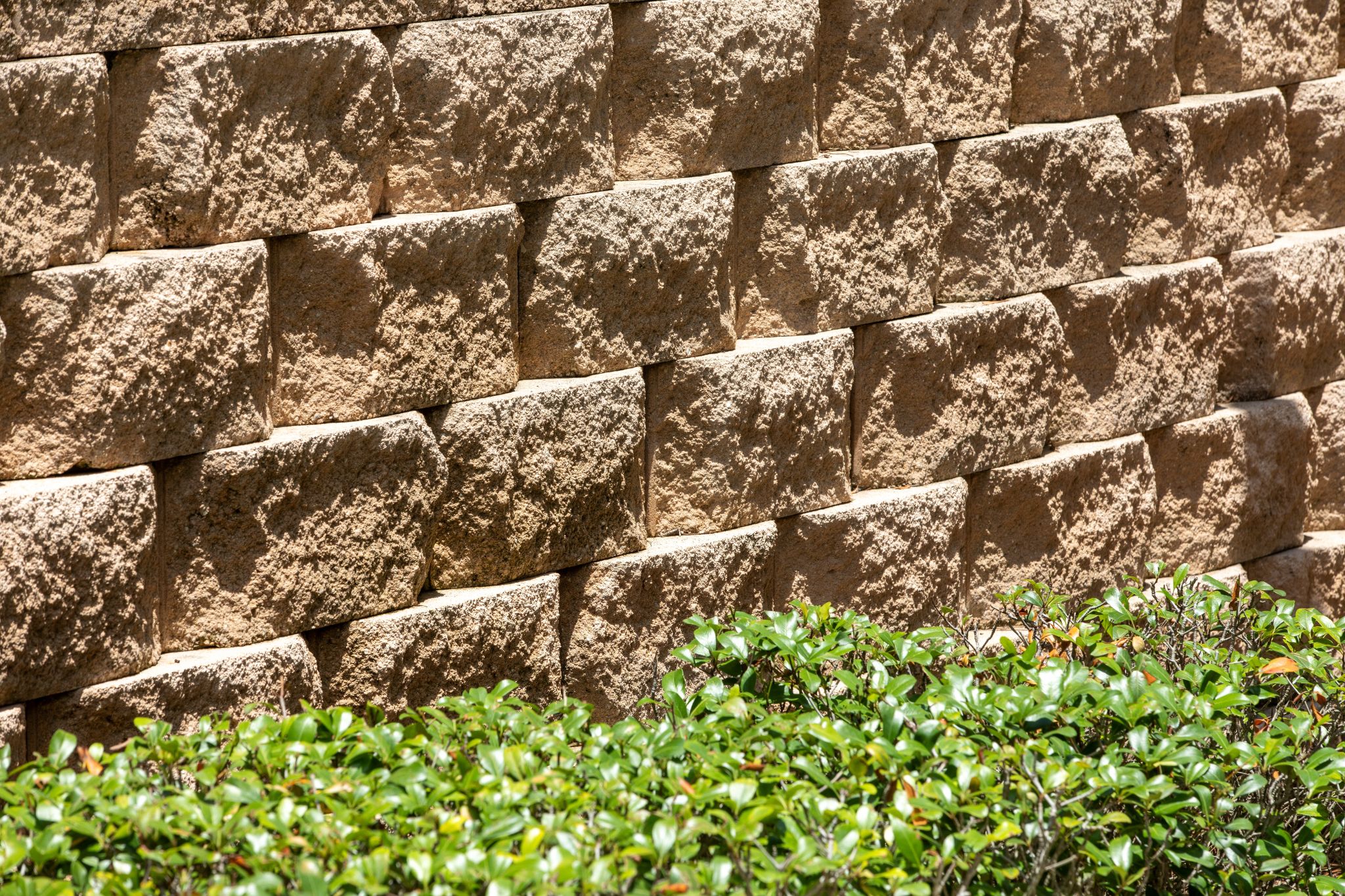 Close-up view of a textured, tan concrete block retaining wall with green shrubbery in the foreground.