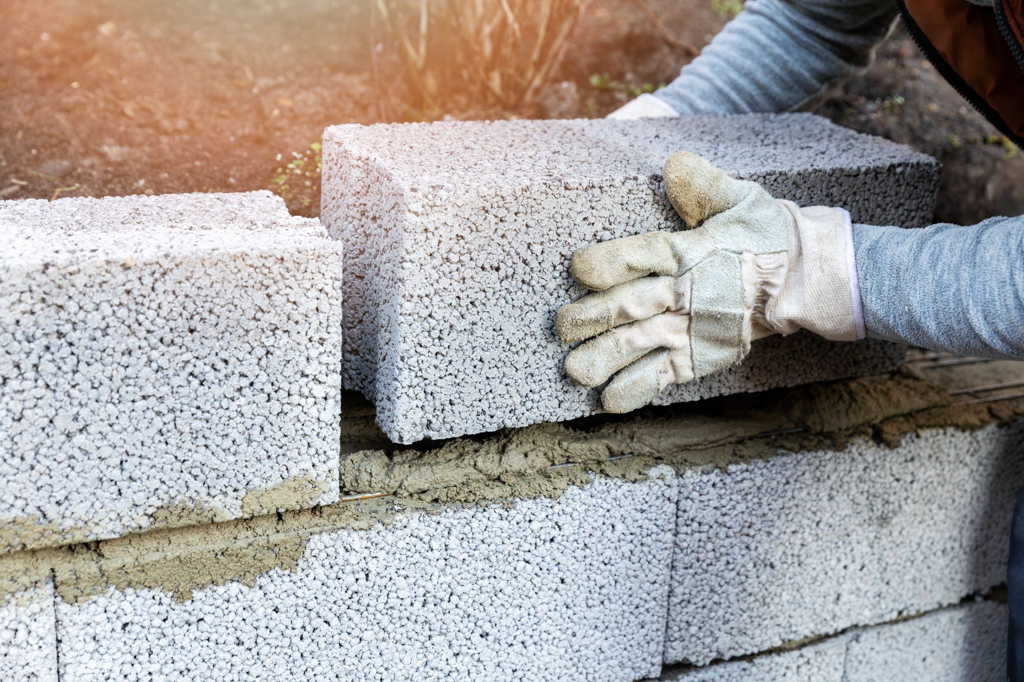 Person wearing work gloves carefully placing a grey concrete block onto mortar while building a wall.