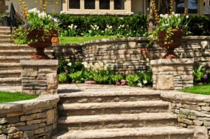 Tiered stone landscape stairs with retaining walls, planted flowers, and urn planters flanking the entrance steps.