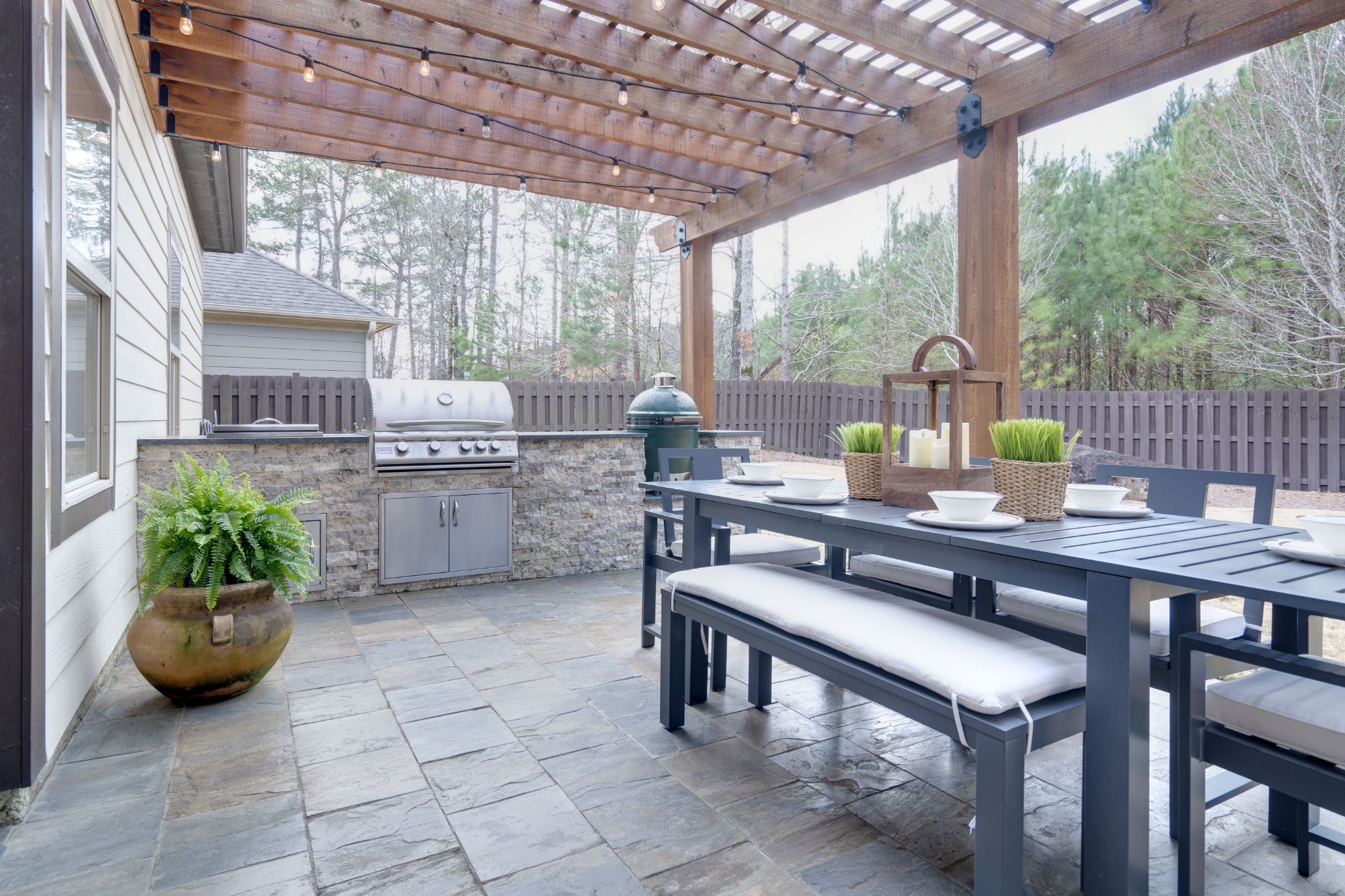 Covered patio with a wooden pergola, outdoor dining table set, string lights, and a stone outdoor kitchen island with a built-in grill.