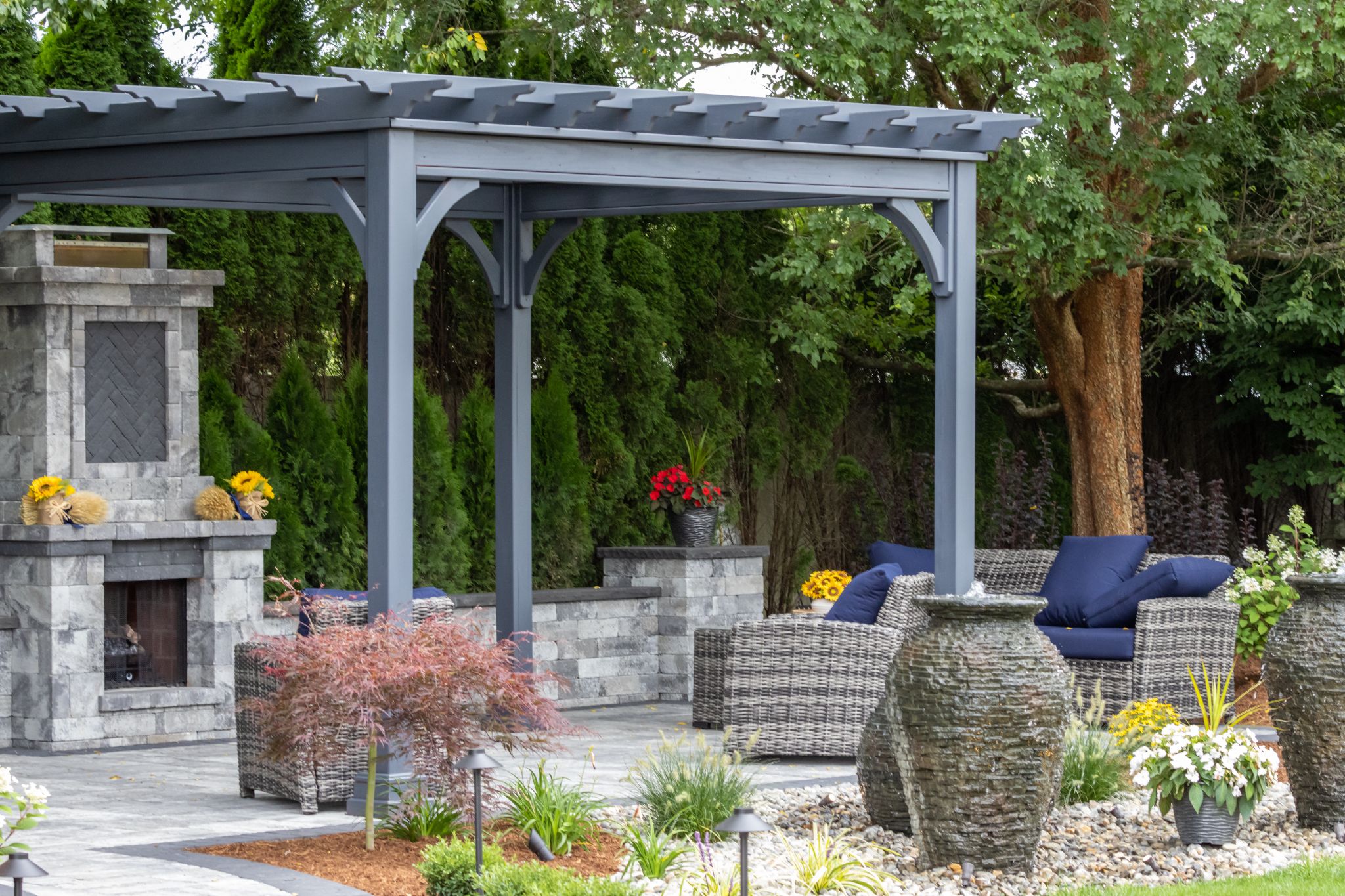 Grey pergola-covered patio with an outdoor stone fireplace, wicker seating, and decorative water urn fountains.