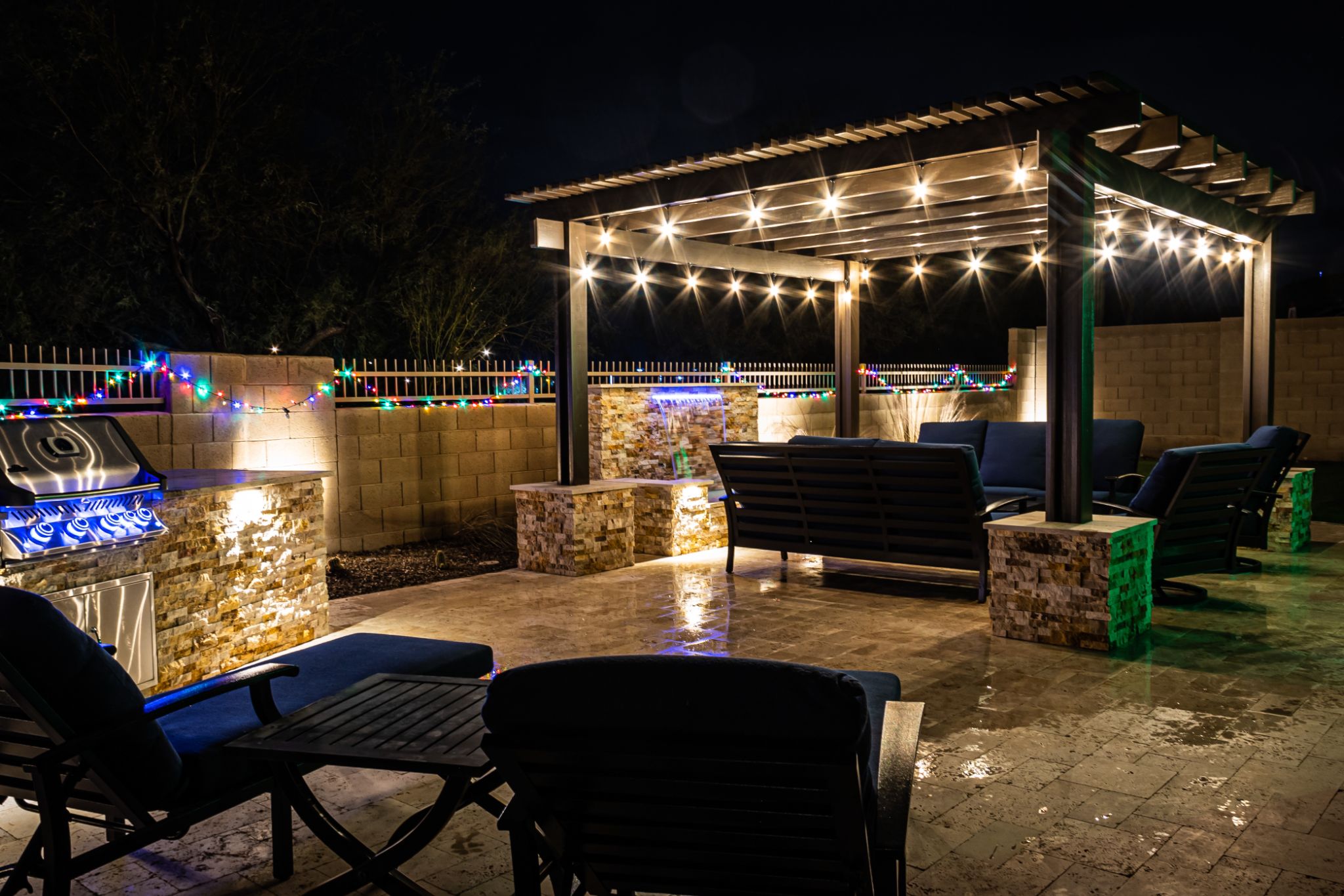 Illuminated backyard patio at night with a pergola, string lights, outdoor seating, built-in stone grill station, and festive fence lights.