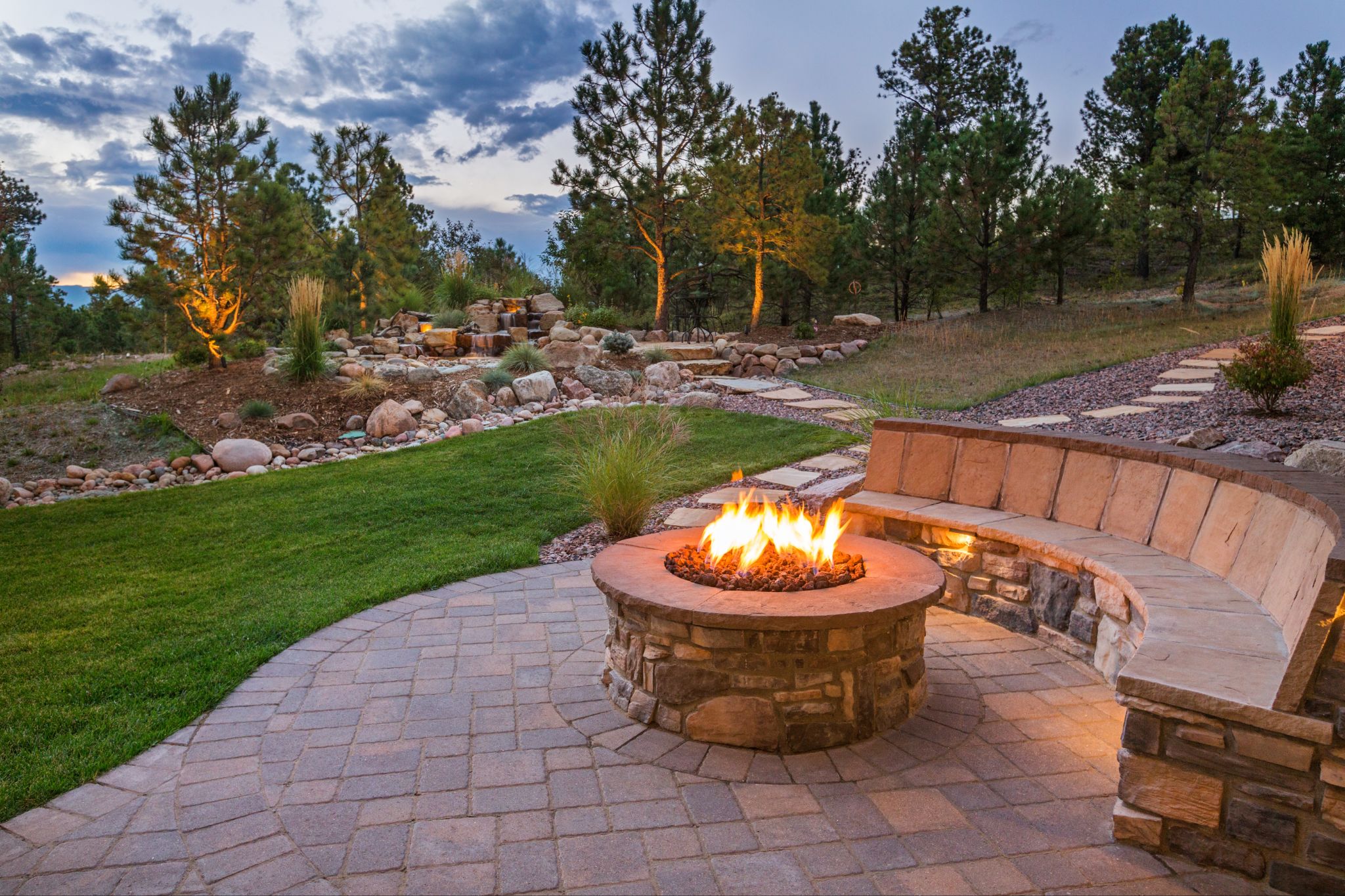 Round stone fire pit with glowing flames and built-in curved stone seating on a paved patio overlooking a landscaped yard at dusk.